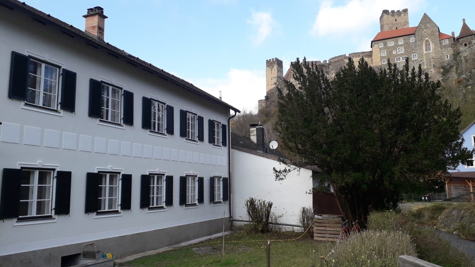 Ein weißes Gebäude mit schwarzen Fensterläden und eine Burg im Hintergrund.