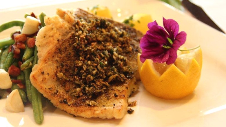 Ein Teller mit gebratenem Fischfilet, dekoriert mit einer Blume und einer Zitronenscheibe, serviert mit grünen Bohnen und Speck.