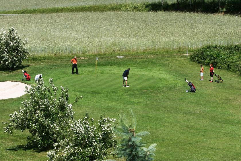 Menschen spielen Golf auf einem grünen Platz mit Bäumen und einem Sandbunker.