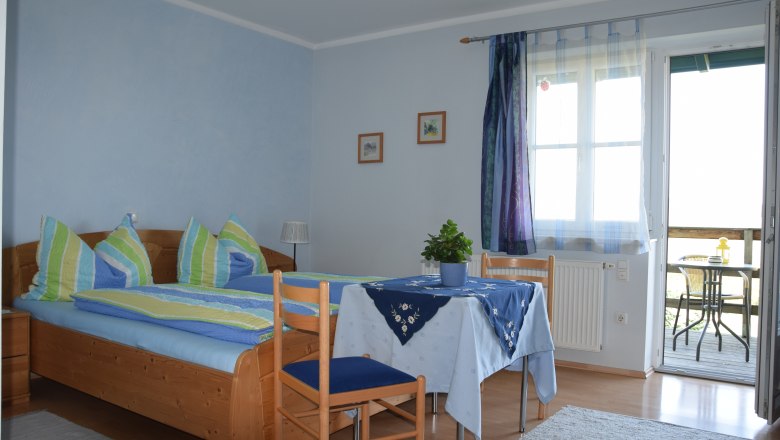Gemütliches Gästezimmer mit Doppelbett, Tisch und Stühlen, blauen Vorhängen und Zugang zu einem Balkon mit Sitzgelegenheit.