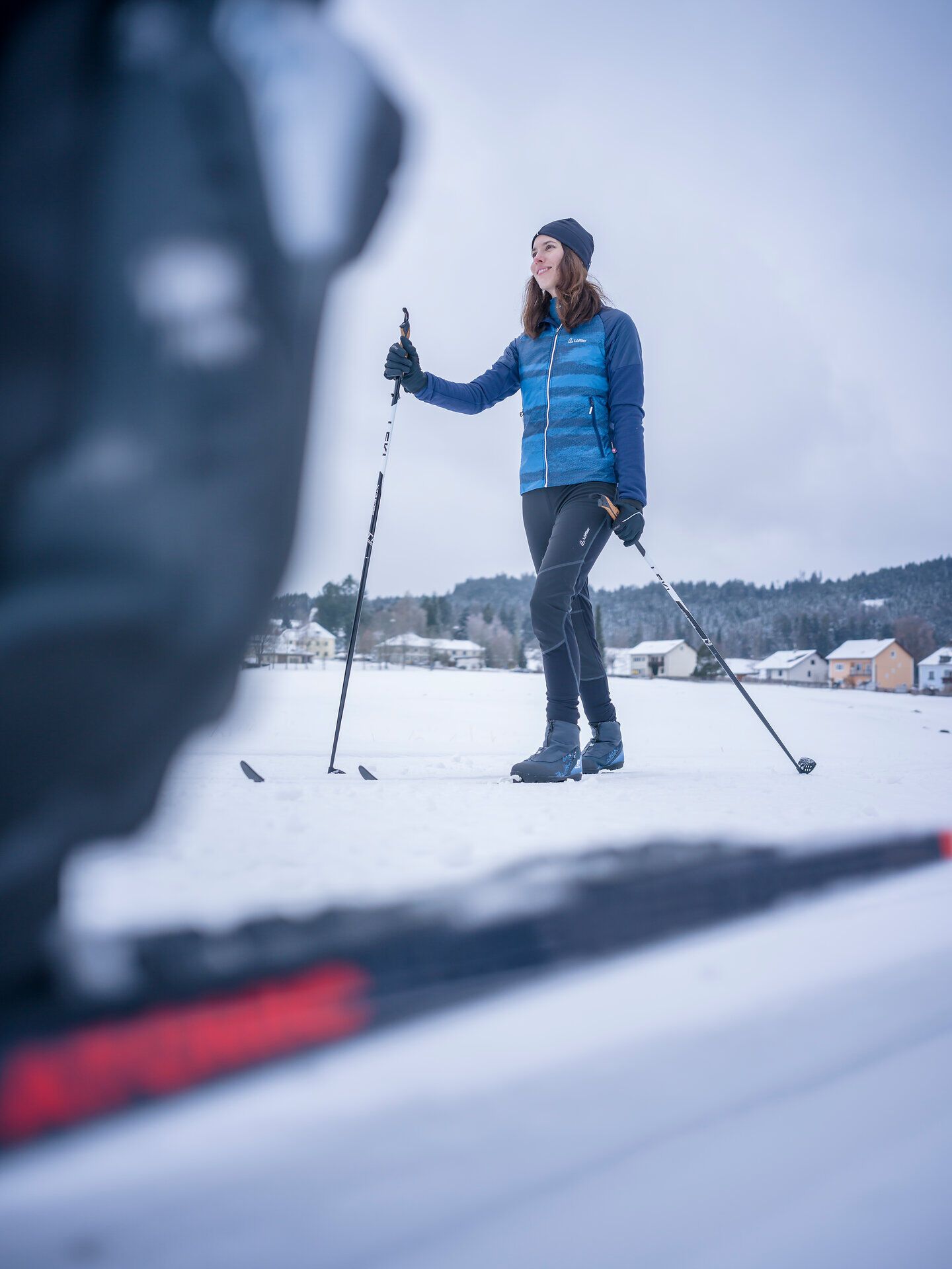 Eine Frau beim Langlaufen auf einer schneebedeckten Fläche mit Häusern im Hintergrund.