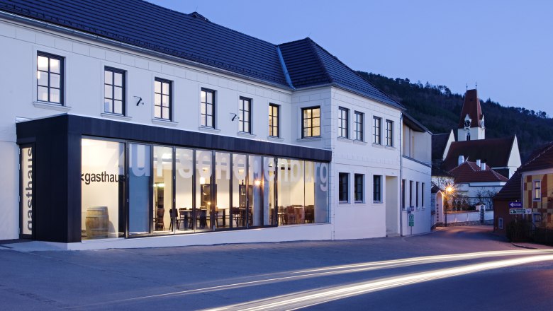 Außenansicht des Gasthofs Hotel Zur Schonenburg bei Dämmerung mit beleuchteten Fenstern und vorbeifahrenden Autos.