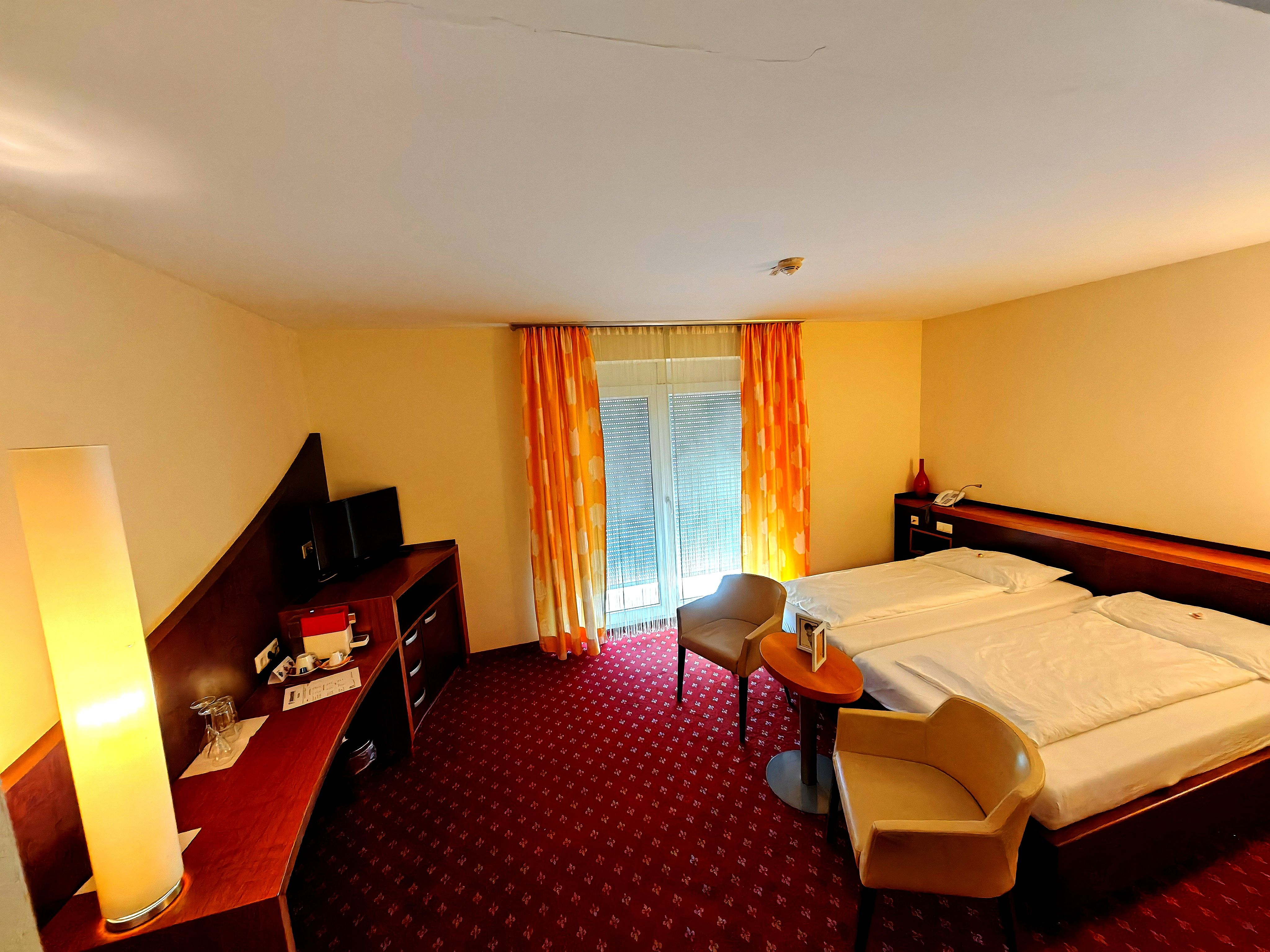 Hotelzimmer mit Doppelbett, zwei Stühlen, einem Tisch, einem Schreibtisch mit Fernseher und einer Stehlampe.