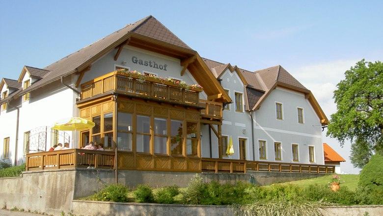 A traditional inn with a wooden veranda and flowers on the balcony, surrounded by green countryside.