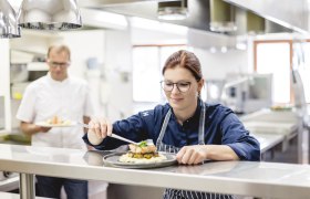 Kuchař v restaurační kuchyni připravuje pokrm, zatímco jeho kolega v pozadí drží jiný pokrm.