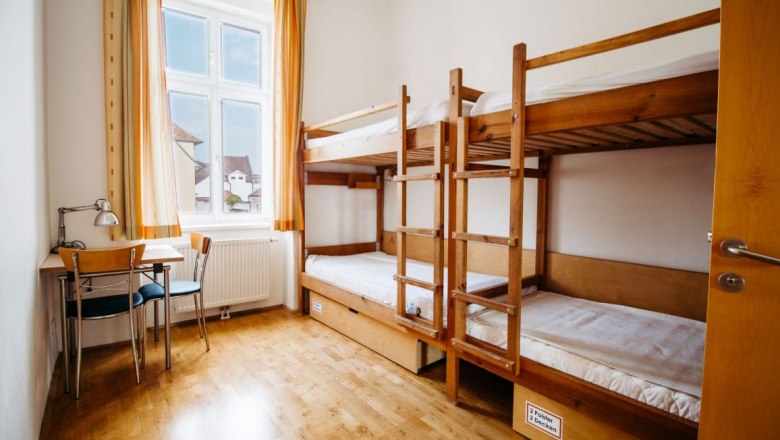 Ein einfaches Zimmer mit Etagenbett, Tisch und Stuhl, helle Vorhänge am Fenster.