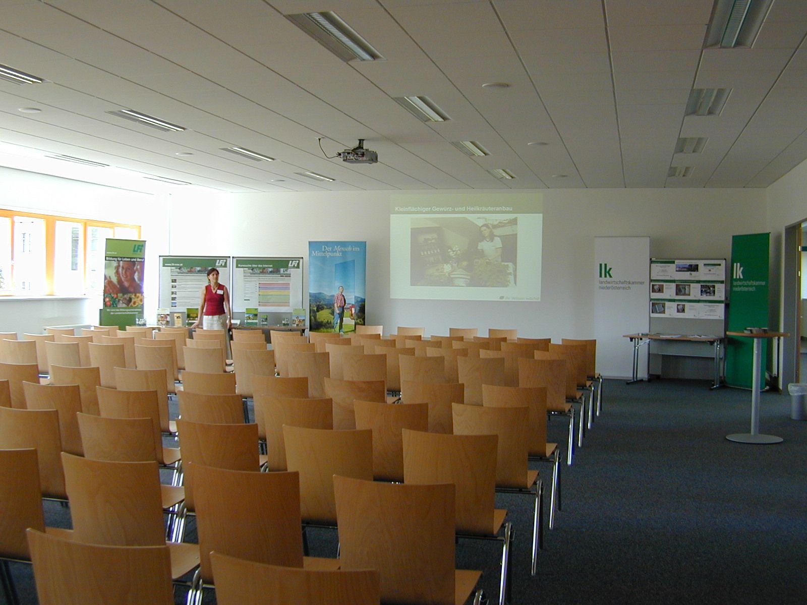 Ein leerer Seminarraum mit Holzstühlen, einer Leinwand und Werbebannern.
