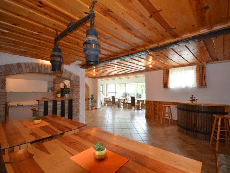 Innenansicht eines rustikalen Gastraums mit Holztischen und -decken, Barbereich und großen Fenstern.