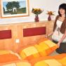 Eine Frau steht in einem Schlafzimmer mit orange-gelber Bettwäsche und Blumenvasen auf dem Kopfteil.