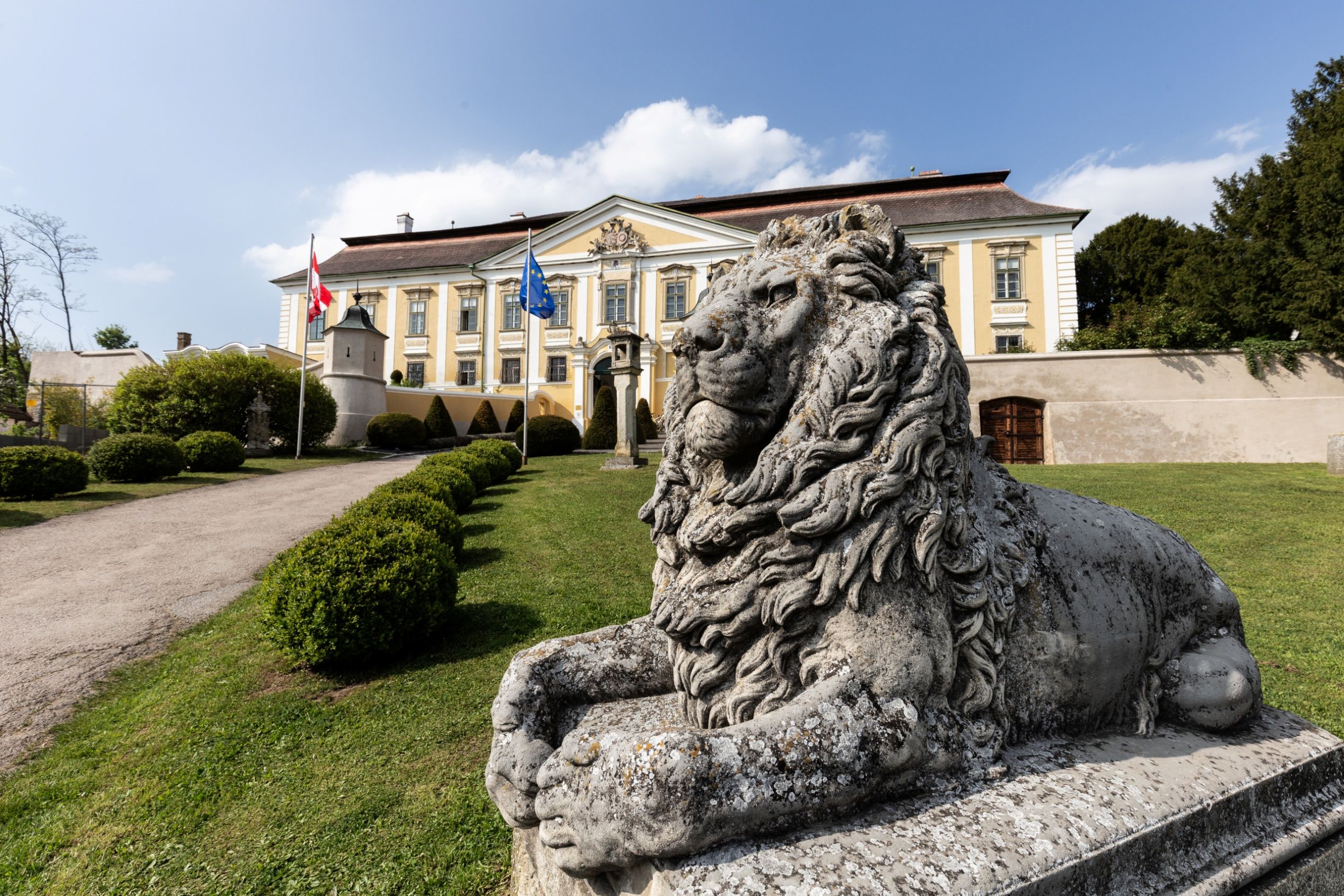 Steinlöwe im Vordergrund vor Schloss Gobelsburg mit österreichischer und EU-Flagge, gepflegtem Garten und barocker Fassade.