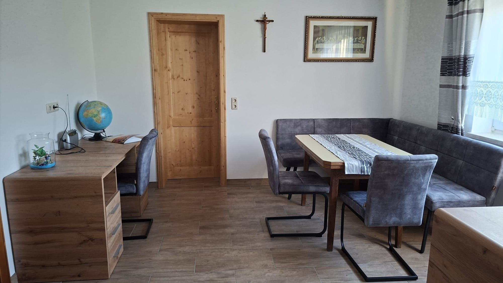 Esszimmer mit Holztisch, grauen Stühlen, einem Schreibtisch mit Globus und einem Kreuz an der Wand.