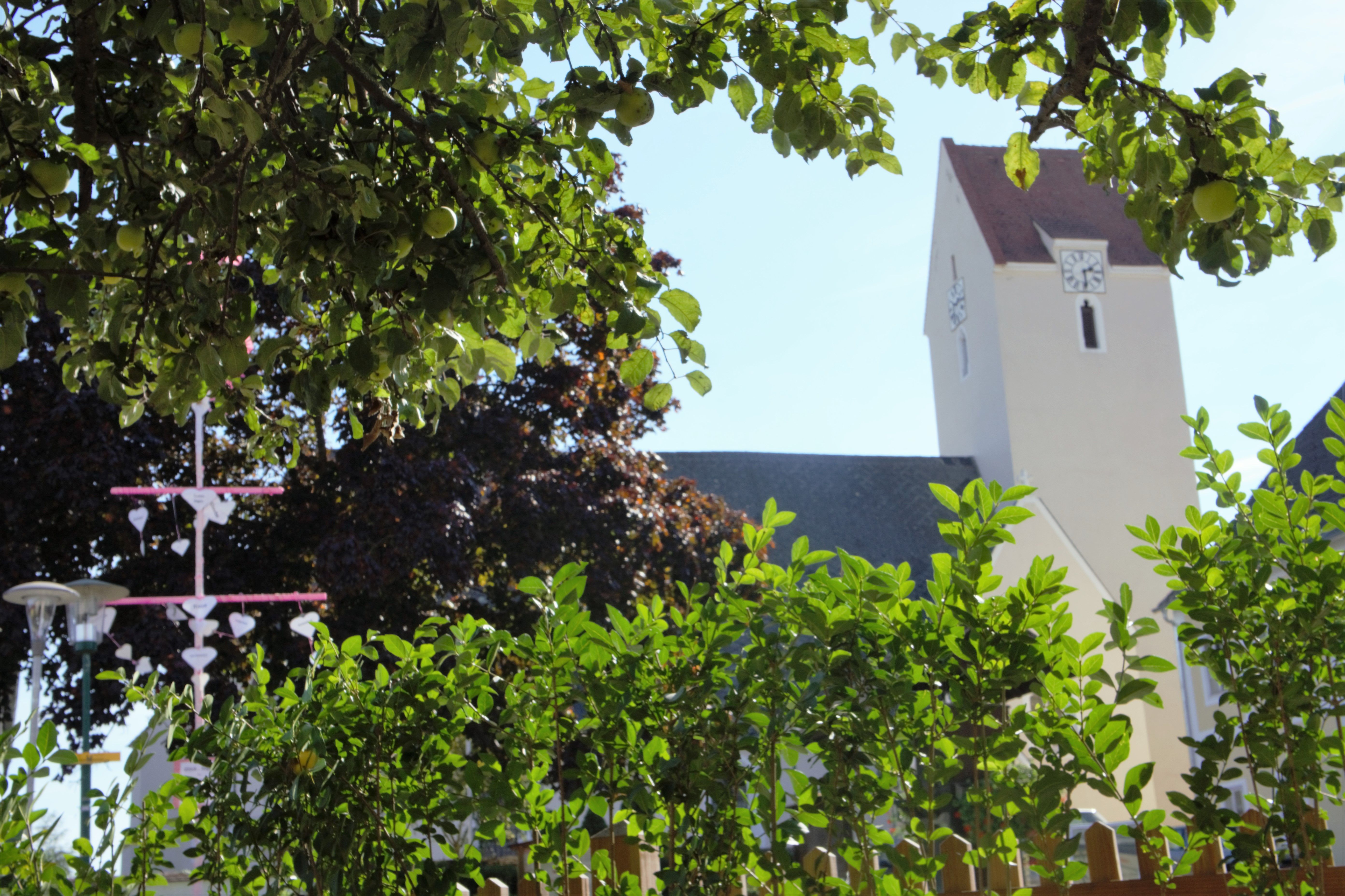 Ein Kirchturm hinter grünen Bäumen und Sträuchern, mit einem dekorativen Element im Vordergrund.