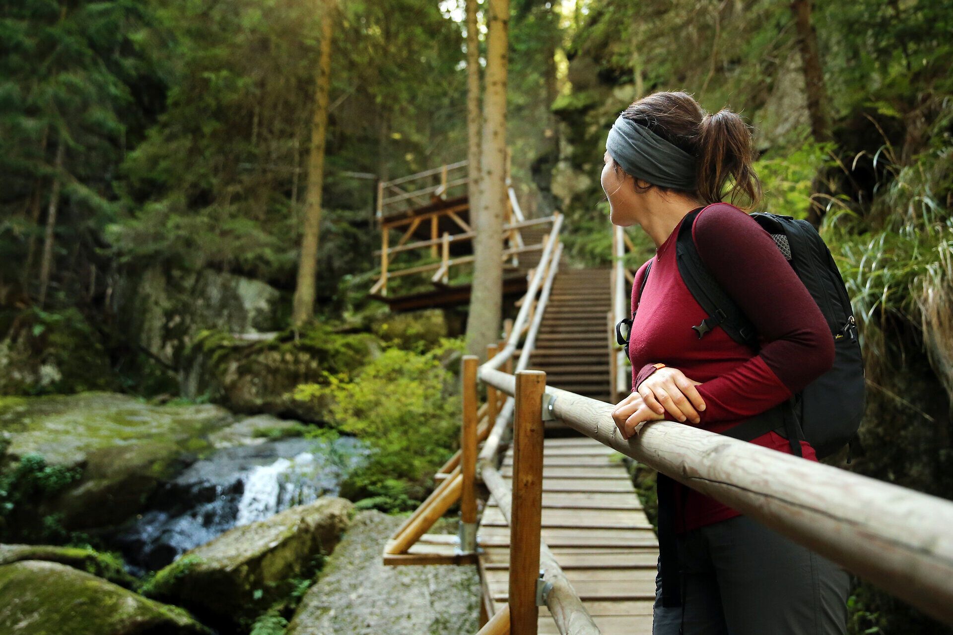 Ein sanfter Wasserfall plätschert über moosbedeckte Steine, während die umgebenden Bäume in sattem Grün erstrahlen. Die Wanderin genießt den Anblick und die frische Luft, die den Sommer in den Bergen so besonders macht.