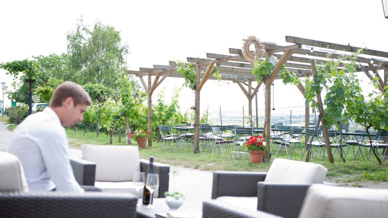 Ein Mann sitzt in einem Gastgarten mit Weinreben und leeren Tischen im Hintergrund.