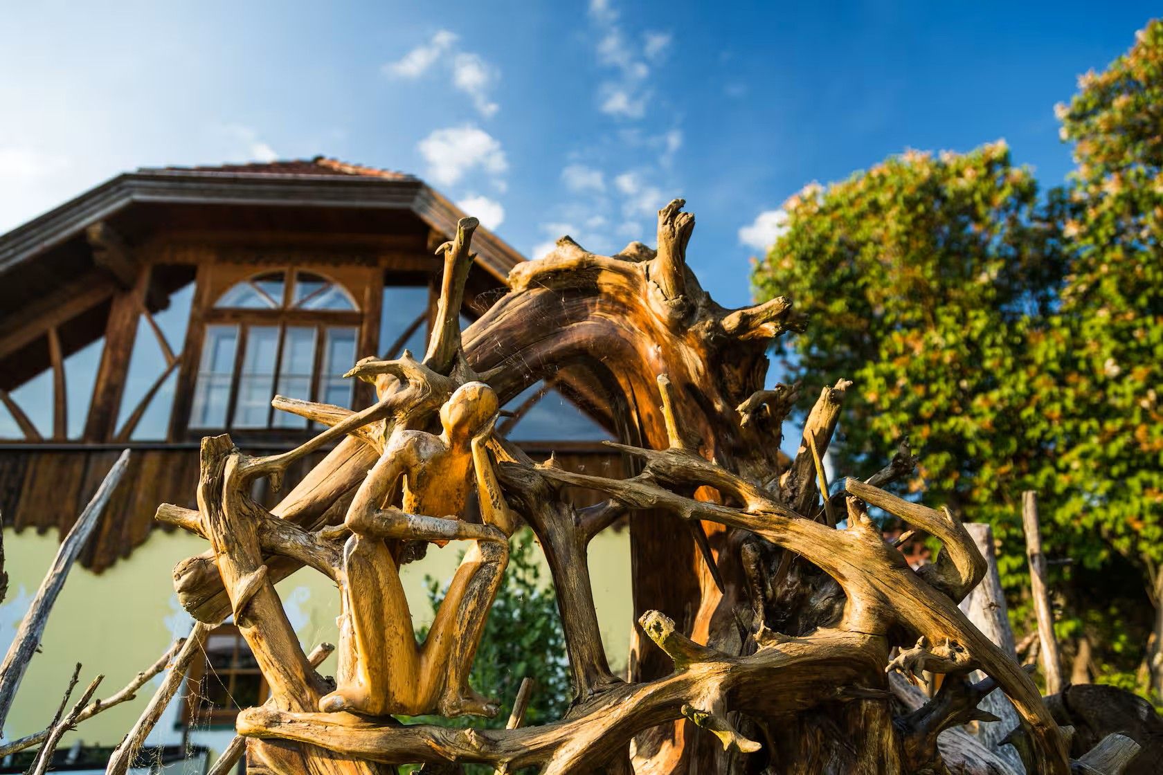 Kunstvolle Holzskulptur vor einem Fachwerkhaus bei blauem Himmel und Bäumen.