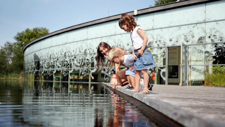 Drei Personen betrachten Wasser an einem Steg vor einem modernen Geb&auml;ude.