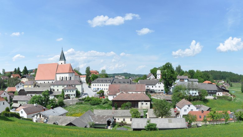 Panorama der Marktgemeinde Sch&ouml;nbach mit Kirche und umliegenden H&auml;usern, umgeben von gr&uuml;nen Wiesen und W&auml;ldern unter blauem Himmel.