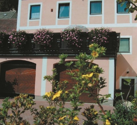 Ein zweistöckiges rosa Gebäude mit blühenden Pflanzen auf dem Balkon und gelben Blumen im Vordergrund.