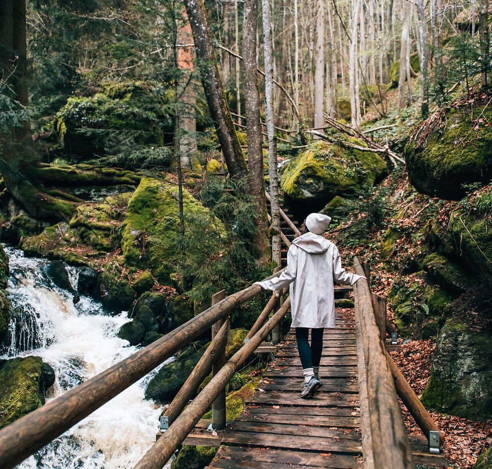 Ein sanfter Wasserfall plätschert über die moosbedeckten Steine, während Wanderer auf der Holzbrücke die Ruhe der Natur genießen. Umgeben von hohen Bäumen und frischem Grün, lädt dieser Ort dazu ein, die Seele baumeln zu lassen und die Schönheit der Umgebung zu erkunden.