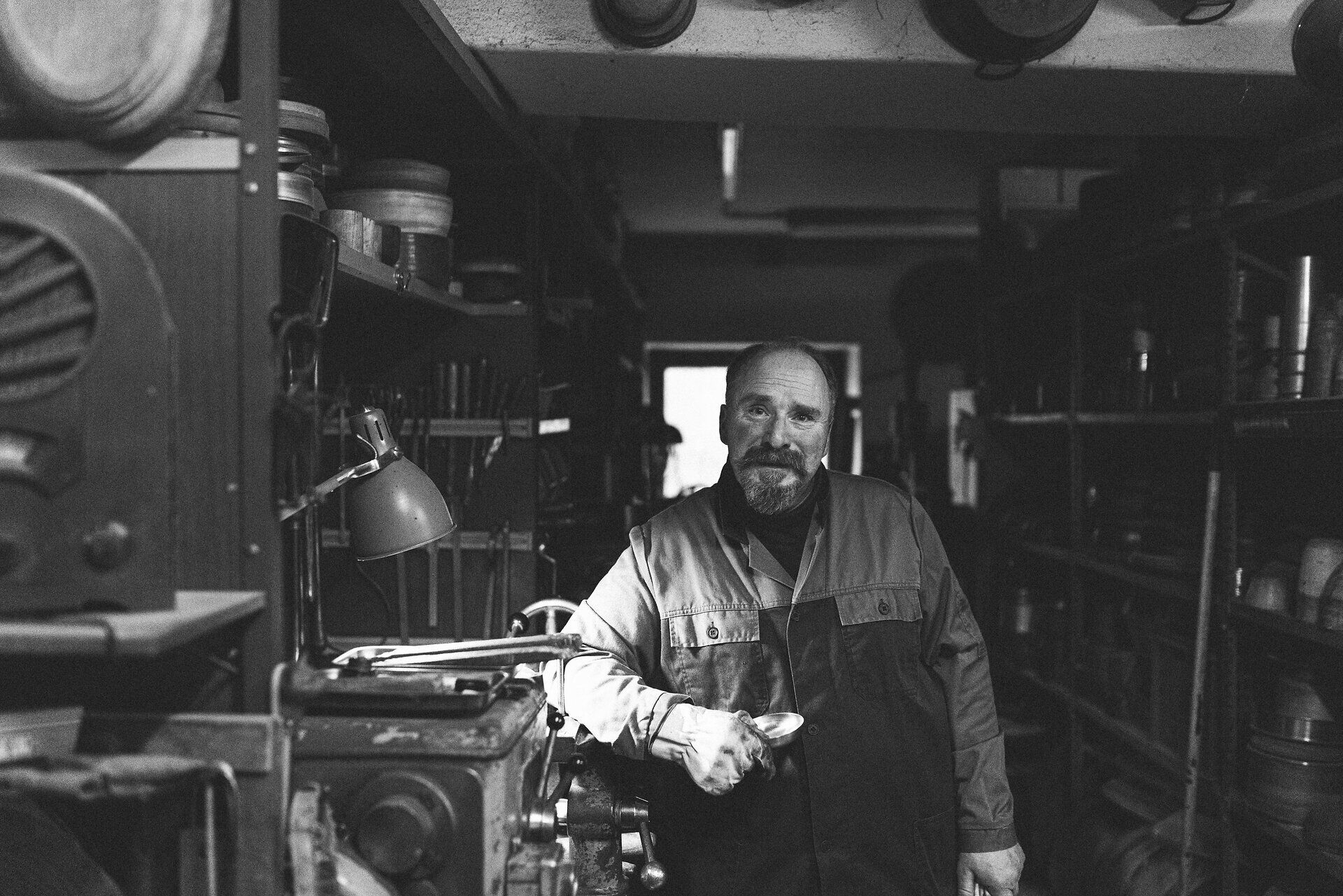 In einer gemütlichen Werkstatt, umgeben von glänzendem Metall und handwerklichem Geschick, steht ein erfahrener Metallverarbeiter. Die Atmosphäre ist geprägt von der Leidenschaft für das Handwerk, während die Kaffeekannen in verschiedenen Formen und Größen auf die Fertigstellung warten.