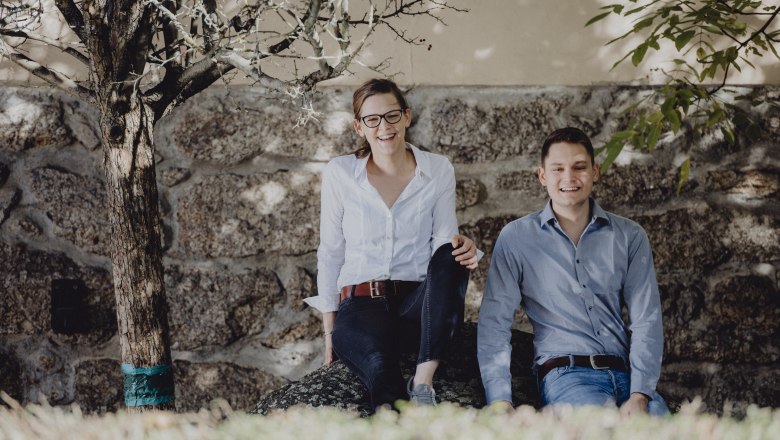 Zwei Personen sitzen lächelnd auf einem Stein vor einer Steinmauer, umgeben von Bäumen.
