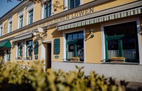 Außenansicht eines gelben Gebäudes mit der Aufschrift 'Zum goldenen Löwen'. Fenster mit Blumenkästen und grünen Markisen.