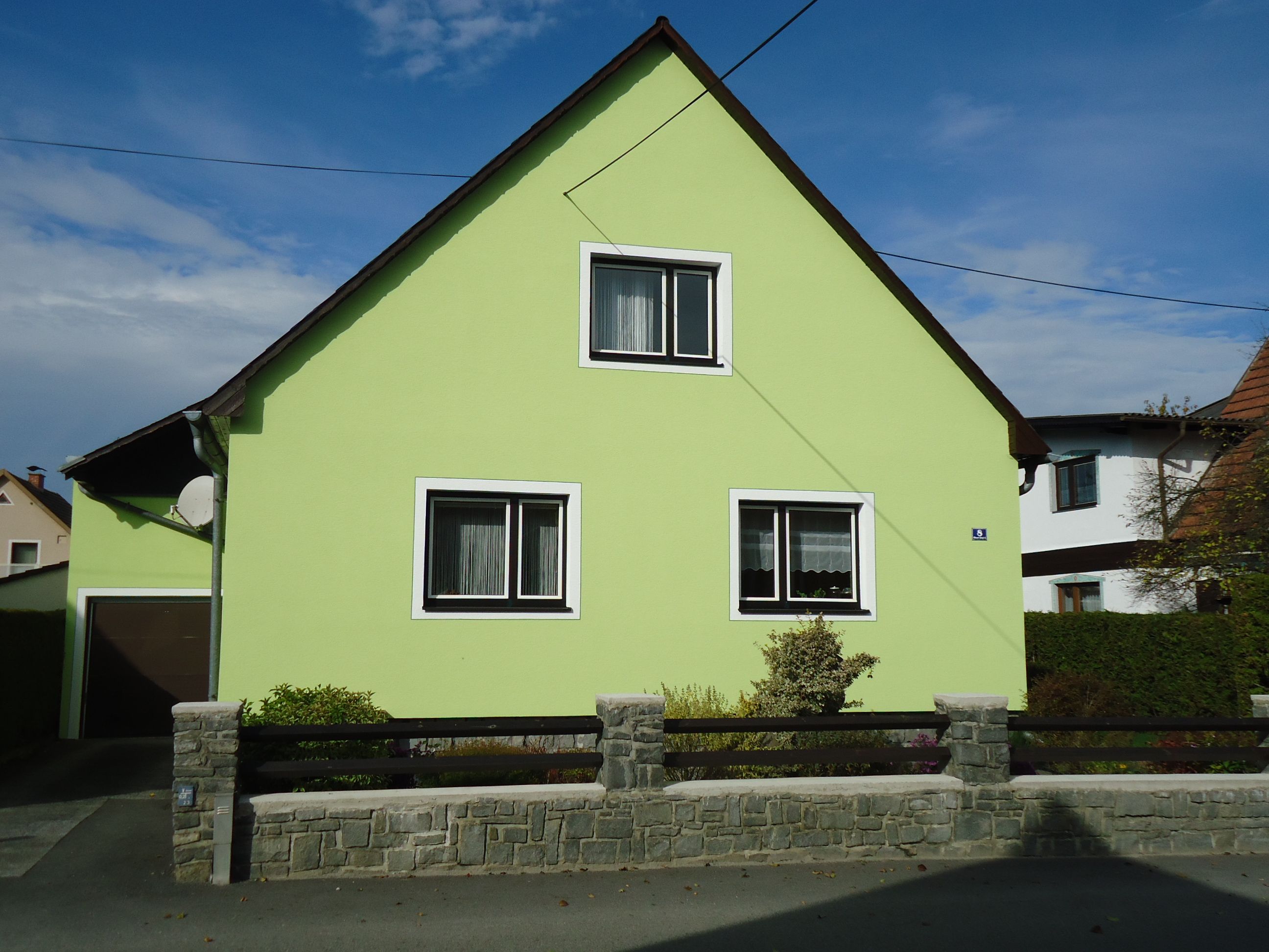 Ein hellgrünes Haus mit Satteldach und zwei Fenstern, umgeben von einem niedrigen Steinzaun.