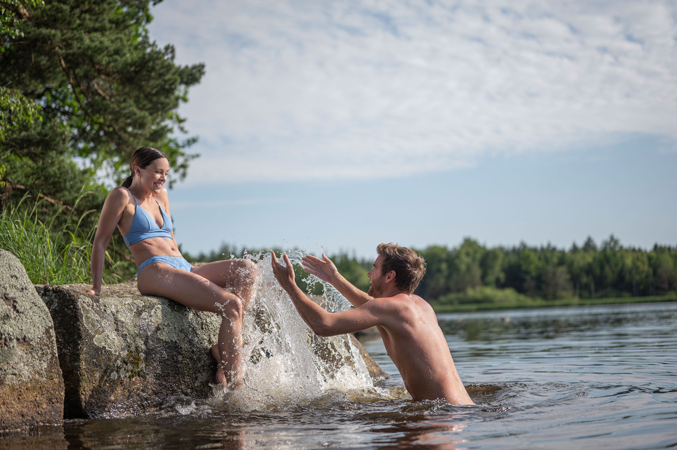 Ein Mann im Wasser spritzt eine Frau an, die auf einem Felsen sitzt.