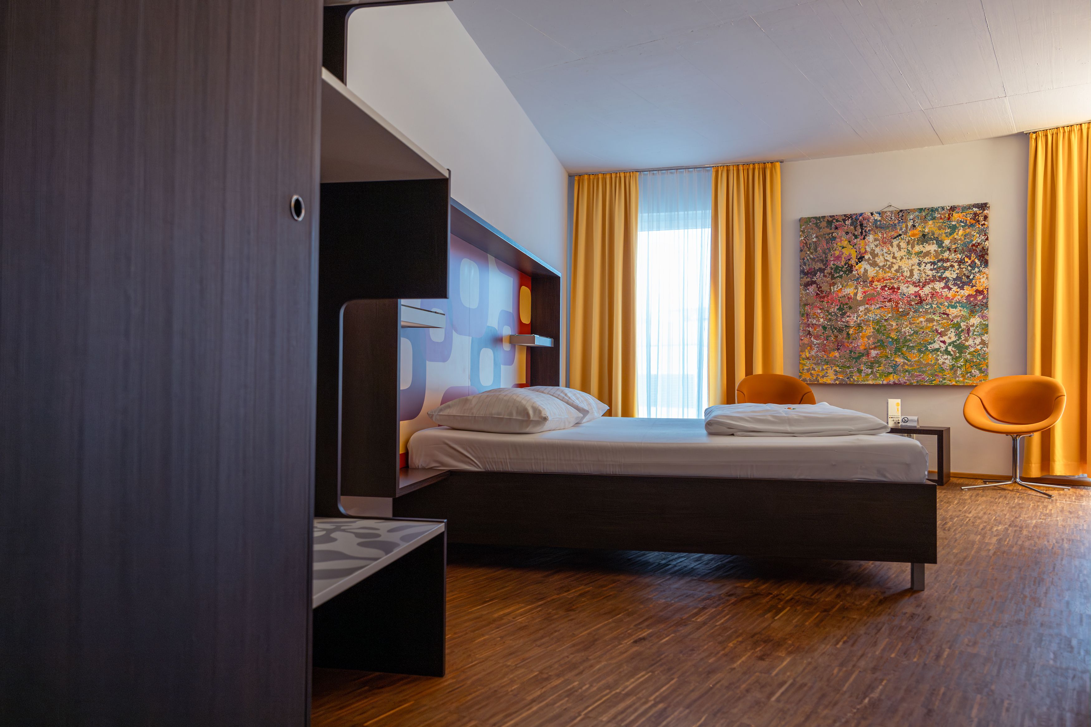 Modernes Hotelzimmer mit Bett, gelben Vorhängen und buntem Gemälde an der Wand.
