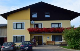 Außenansicht eines gelben Hauses mit Balkon voller roter Blumen und geparkten Autos davor.