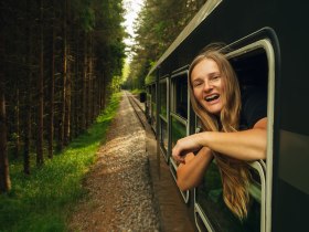 Eine l&auml;chelnde Frau lehnt sich aus dem Fenster eines fahrenden Zuges, der durch einen dichten Wald im Waldviertel f&auml;hrt.