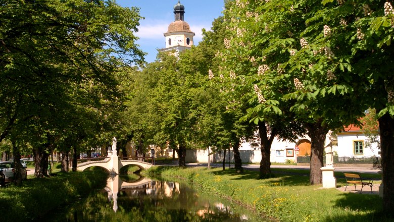 Ein idyllischer Park mit einem kleinen Fluss, einer Brücke und einem Kirchturm im Hintergrund.