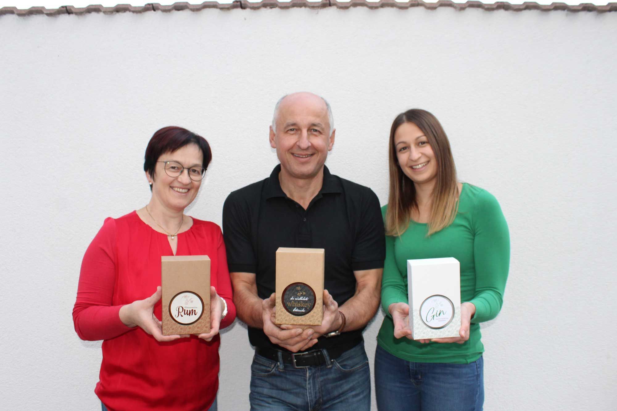 Drei Personen halten Geschenkboxen mit Rum, Whiskey und Gin vor einer weißen Wand.