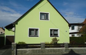 Ein hellgrünes Haus mit Satteldach und zwei Fenstern, umgeben von einem niedrigen Steinzaun.