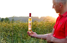 Ein Mann in rotem Hemd hält eine Flasche Dinkelbrand in einem Getreidefeld bei Sonnenuntergang.