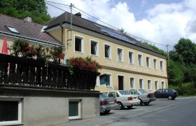 Gelbes Gebäude mit der Aufschrift 'Gasthof zum guten Tropfen Josef Rucker', Autos parken davor, blauer Himmel im Hintergrund.