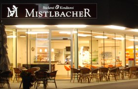 Au&szlig;enansicht eines Caf&eacute;s mit dem Namen Mistelbacher, beleuchtet und mit Sitzgelegenheiten im Freien.