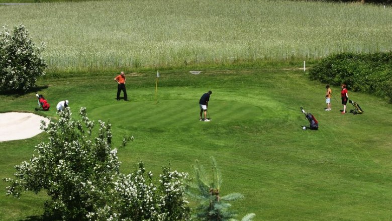 Menschen spielen Golf auf einem grünen Platz mit Bäumen und einem Sandbunker.