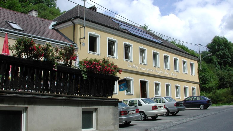 Gelbes Gebäude mit der Aufschrift 'Gasthof zum guten Tropfen Josef Rucker', Autos parken davor, blauer Himmel im Hintergrund.