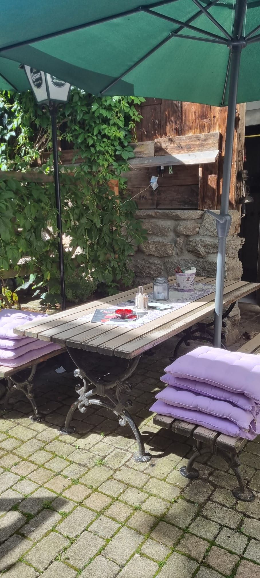 Ein rustikaler Holztisch mit Bänken und lila Kissen unter einem grünen Sonnenschirm im Freien.