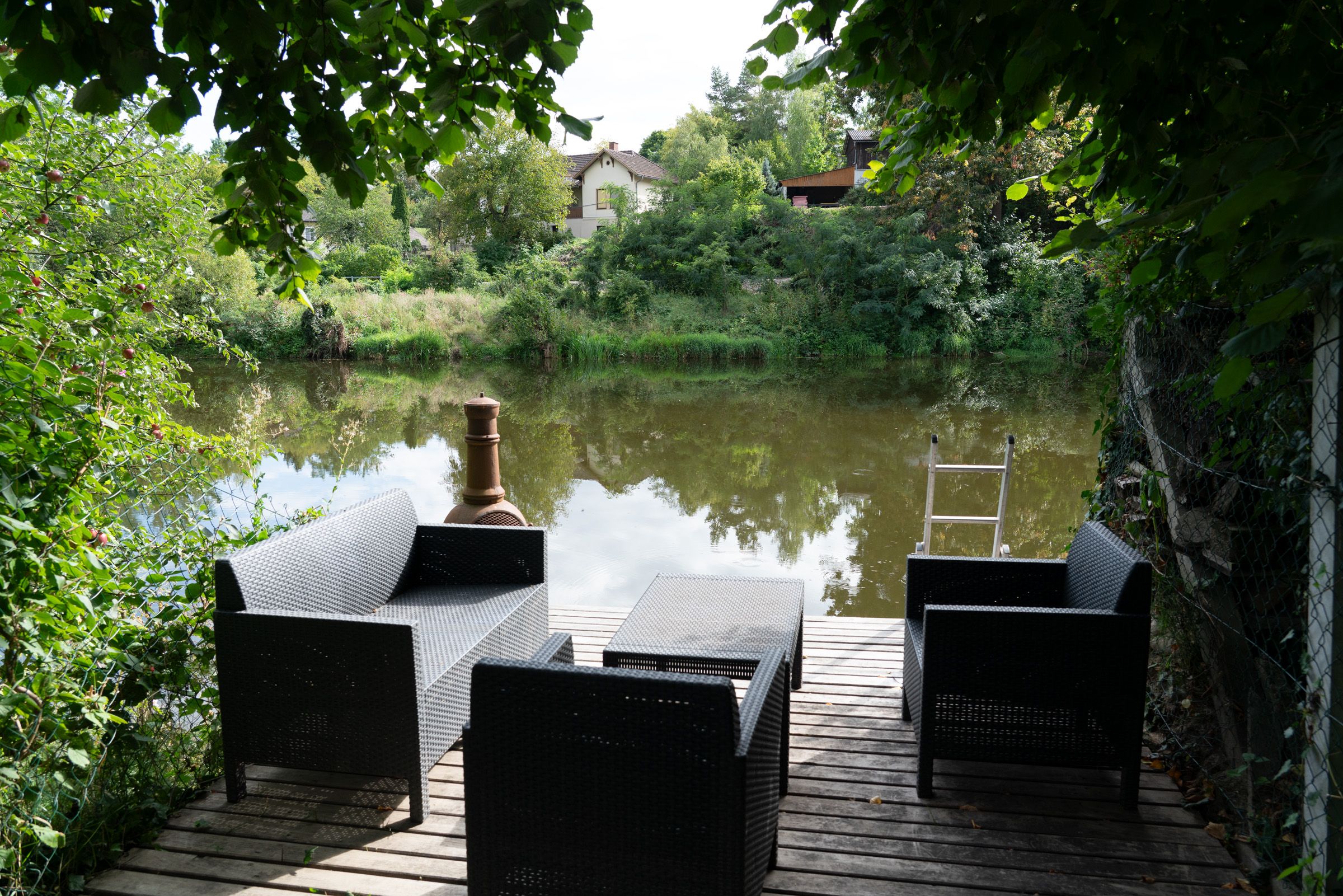 Ein Holzsteg mit schwarzen Gartenmöbeln am Flussufer, umgeben von grüner Vegetation und einem Haus im Hintergrund.