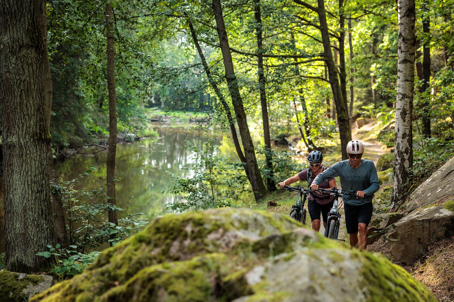 Inmitten der üppigen Wälder und sanften Hügel wandern zwei Radfahrer entlang eines malerischen Pfades, der von hohen Bäumen gesäumt ist. Die ruhige Atmosphäre und das sanfte Plätschern des Wassers im Hintergrund laden dazu ein, die Schönheit der Natur in vollen Zügen zu genießen.