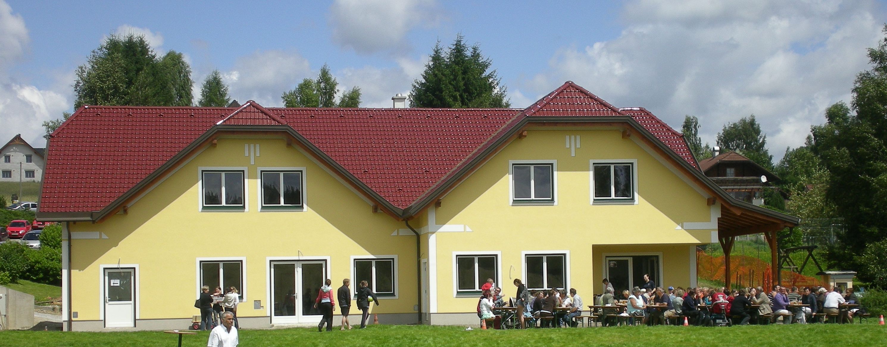 Gelbes Gebäude mit rotem Dach, Menschen sitzen draußen an Tischen.