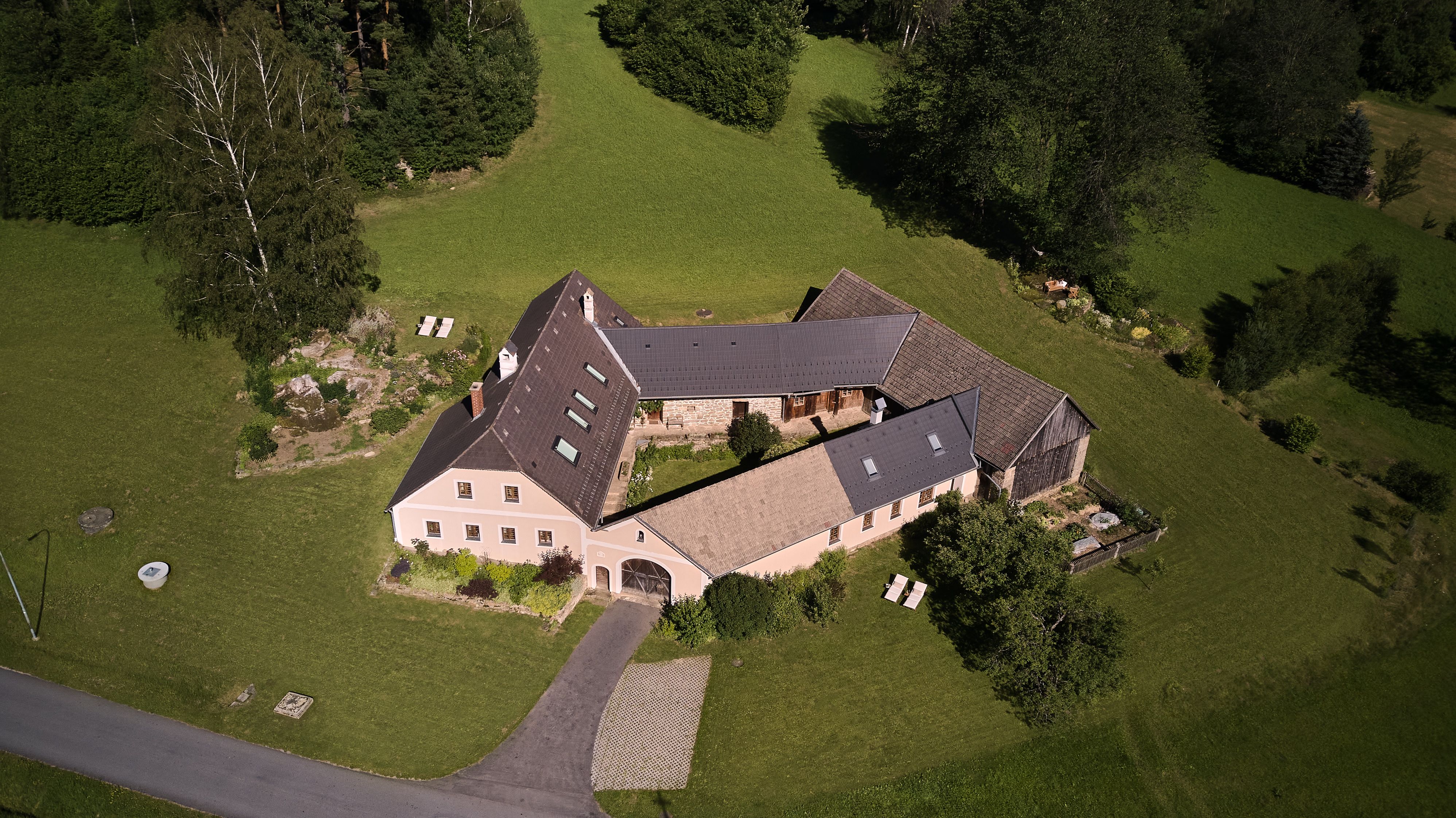 Luftaufnahme eines großen, L-förmigen Gebäudekomplexes auf einer grünen Wiese, umgeben von Bäumen.