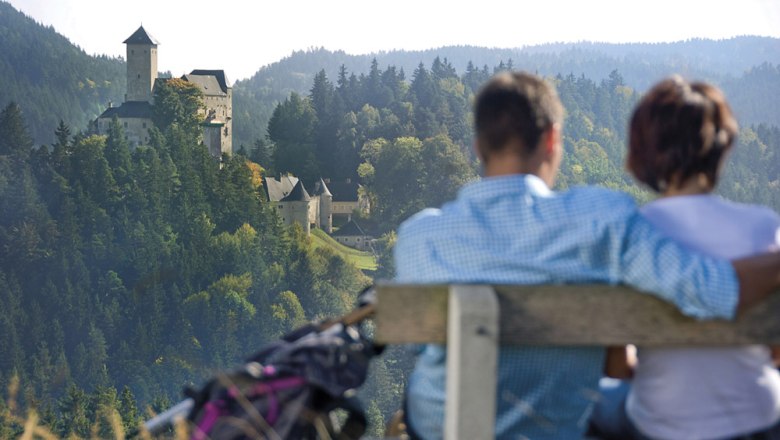 Ein Paar sitzt auf einer Bank mit Blick auf eine Burg in einer bewaldeten Landschaft.