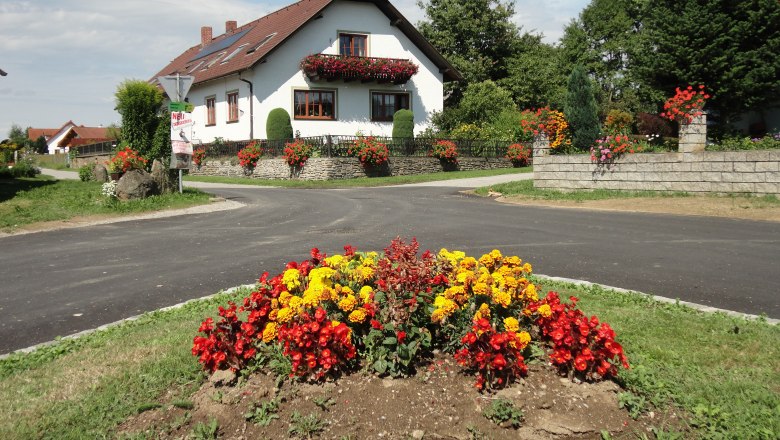 Ein wei&szlig;es Haus mit roten Dachziegeln und Blumenschmuck, umgeben von bunten Blumenbeeten und gr&uuml;ner Landschaft.