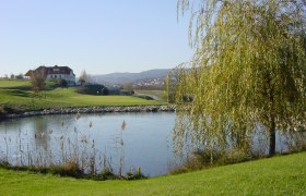 Landschaft mit Golfplatz, Teich und Gebäude im Hintergrund.