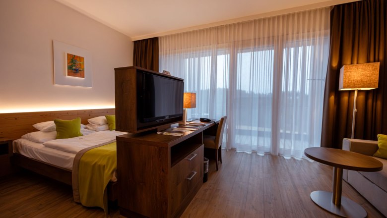 Modernes Hotelzimmer mit Doppelbett, Fernseher, Schreibtisch und großen Fenstern mit Vorhängen.