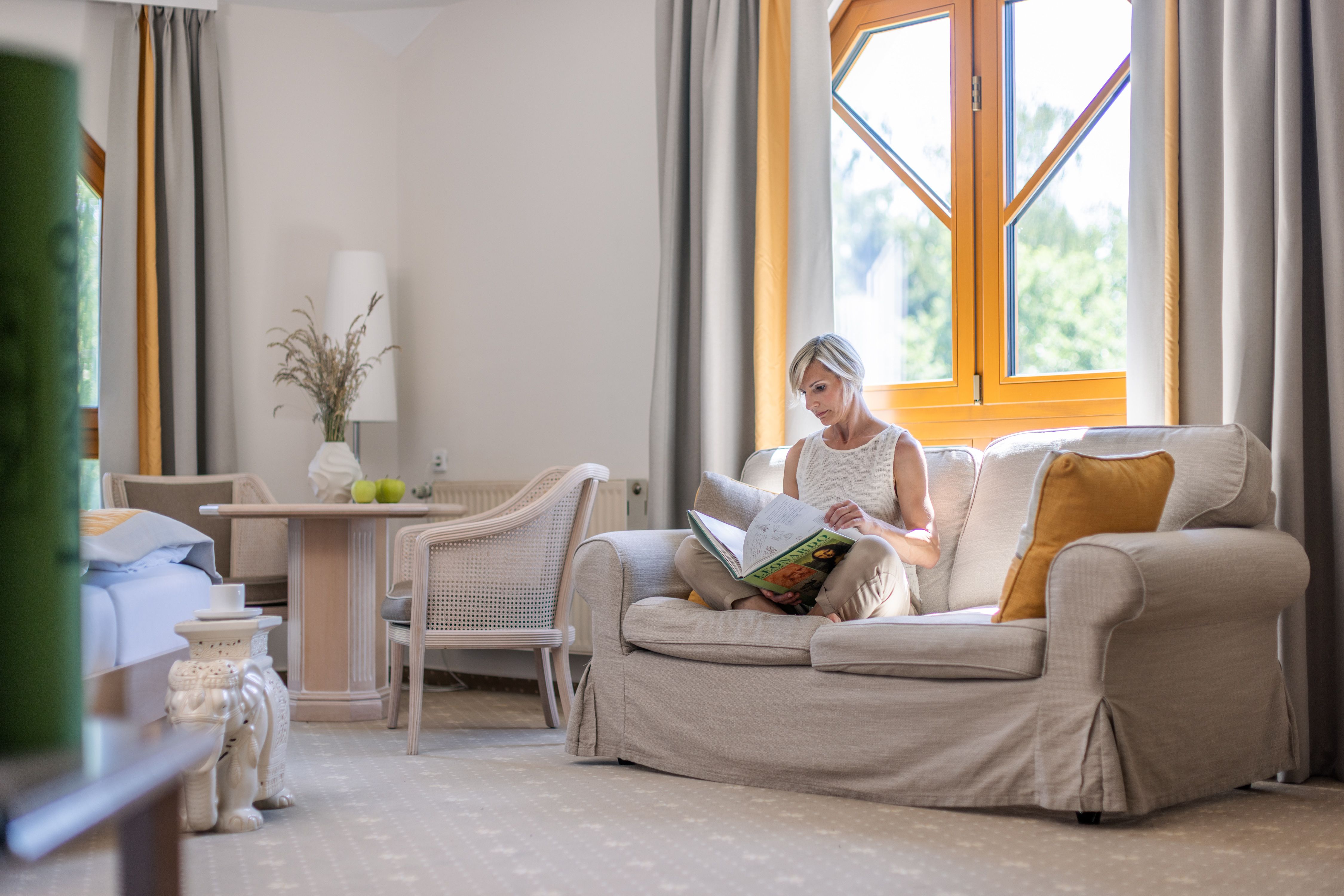 Eine Frau sitzt auf einem Sofa in einem hellen Raum und liest ein Buch. Neben ihr steht ein Tisch mit zwei grünen Äpfeln und einer Vase mit Gräsern.