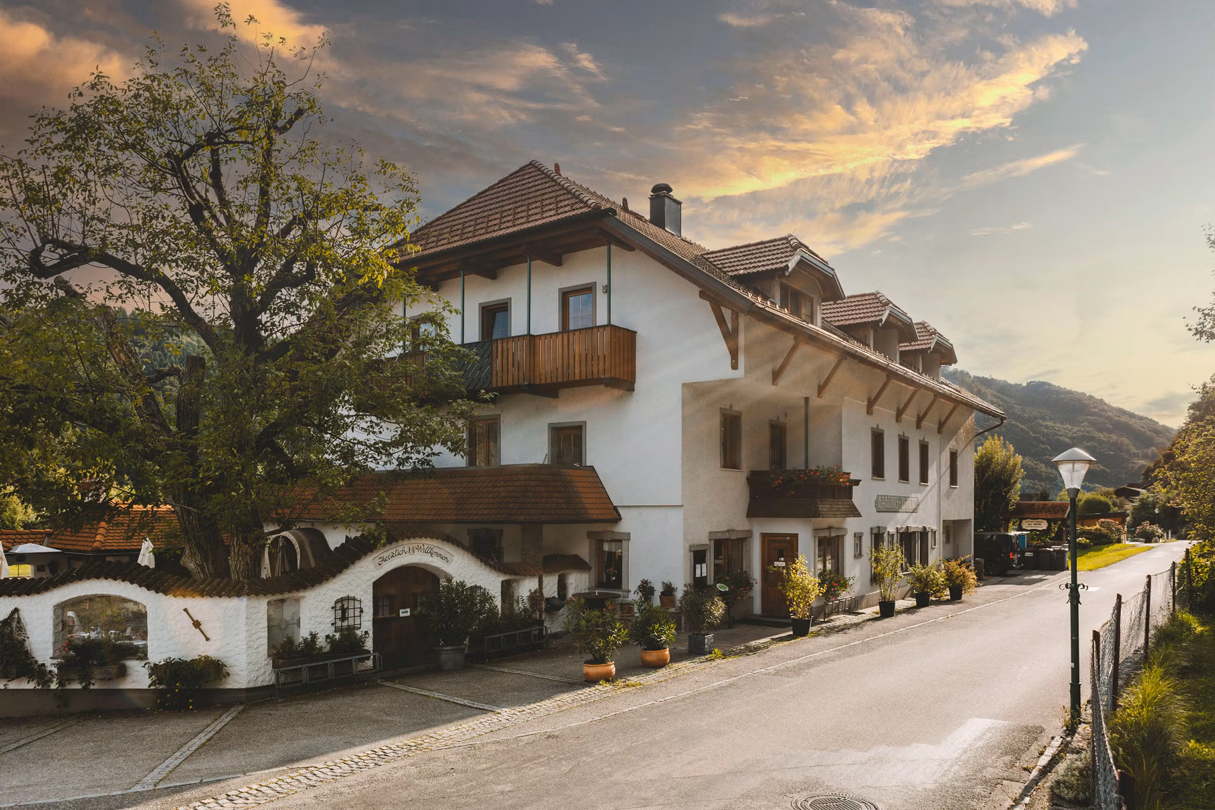 Ein traditionelles Wirtshaus mit weißen Wänden und roten Ziegeldächern, umgeben von Bäumen und einer ruhigen Straße.